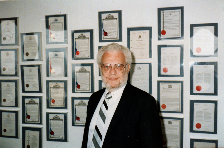 Robert S. Block is wearing a black suit and black and white stripped tie, as he stands in front of a wall patents granted to him for various endeavors.